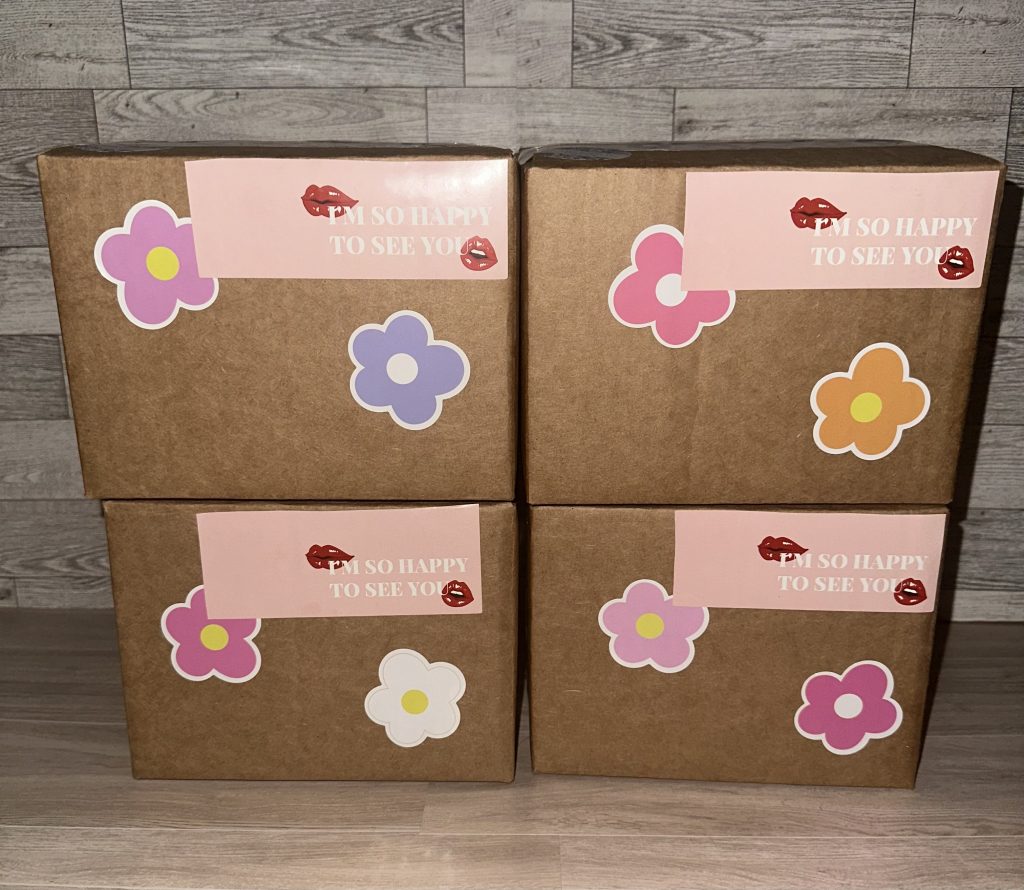 Four packaged boxes styled on a clean, aesthetically pleasing background, each decorated with two flower stickers and a I’m so happy to see you sticker with lips next to the words.